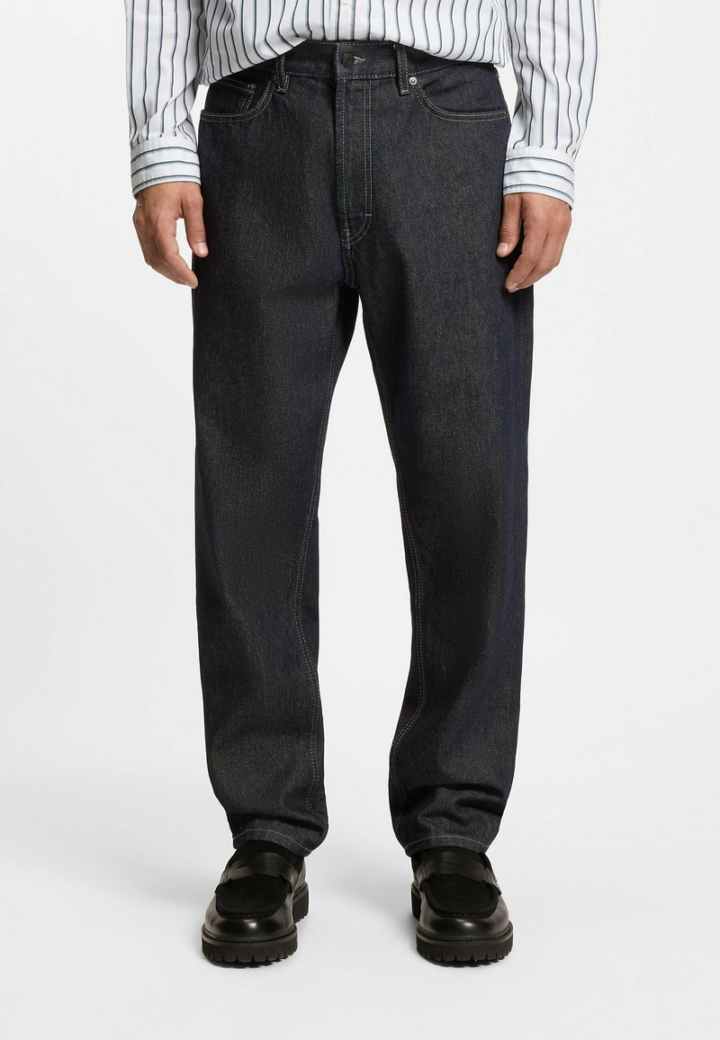 Jeans in denim blu scuro con taglio a gamba dritta, caratterizzati da tasche frontali e dettagli di cucitura sottili. Indossati con mocassini neri e una camicia a righe.