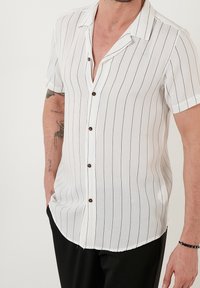 Muž oblečený v bílém krátkém rukávu košili s tenkými vertikálními černými pruhy a černých kalhotách, s jednou rukou v kapse a viditelnými tetováními na pažích.