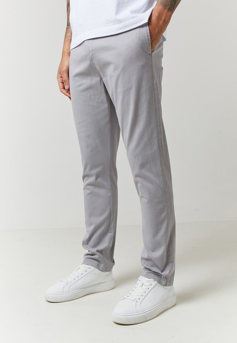 Szürke pamut nadrág, slim fit szabással, oldalsó zsebekkel és bokáig érő szegéllyel, fehér sportcipőkkel egy semleges hátteren.