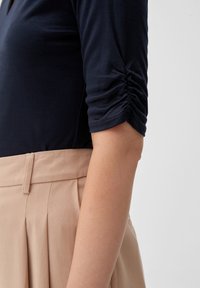 Läheltä otettu kuva henkilön käsivarresta, joka on pukeutunut tummansiniseen rypytettyyn hihapaitaan, joka on työnnetty korkeavyötäröisiin vaaleanbeigeihin laskettuihin housuihin, yksinkertaista taustaa vasten.