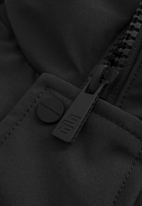 Close-up van zwart stof met een grote zwarte rits en een ronde knop met de tekst "BLACK ENNIE" erop gestikt.