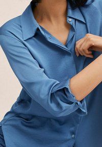 Femme portant une chemise bleue à manches longues retroussées, bras croisés, sur un fond neutre.