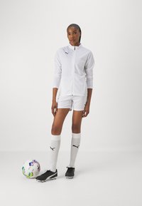 White athletic jacket with zip, matching shorts, black soccer cleats, and white knee-high socks with black logo. Colorful soccer ball at feet.