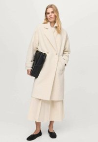 Abrigo de textura cremosa con diseño de doble botonadura, ajuste holgado, combinado con una falda plisada crema y zapatos negros sin cordones. Lleva un bolso negro.