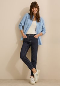 Chemise boutonnée bleu clair par-dessus un débardeur blanc, associée à un jean en denim foncé et des baskets blanches. L'ensemble a une coupe et un style décontractés.