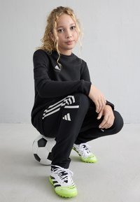 Young person with curly blonde hair squatting on a soccer ball, wearing black Adidas tracksuit and white-green cleats against a plain gray wall.