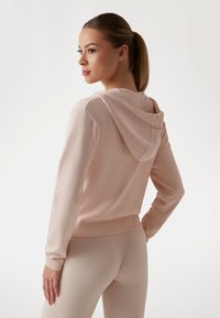 Lyserød ribbet hoodie set fra ryggen, med en afslappet pasform, lange ærmer og hætte med snoretræk, parret med matchende ribbet leggings.