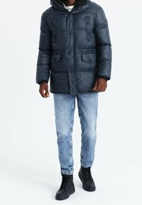 Giacca a vento imbottita blu navy con cappuccio, caratterizzata da due tasche frontali, bottoni a pressione e tessuto strutturato. Abbinata a jeans azzurri e stivali neri.