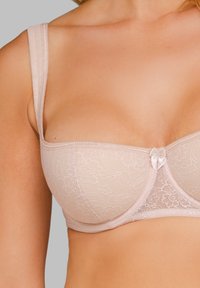 Koronkowy biustonosz usztywniany w jasno-beżowym kolorze z cienkimi ramiączkami, ozdobiony satynową kokardką na środku oraz kwiatowym wzorem na całym materiale.