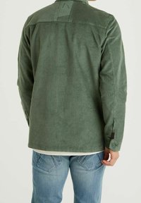 Vihreä samettipaita pystysuorin raidoin, napilliset hihansuut ja selässä olkasauma. Päällä vaaleansiniset farkut.