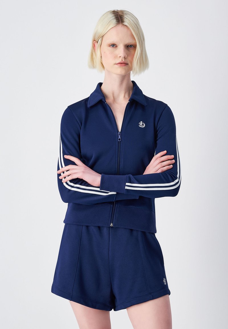 Giacca da tuta con zip blu navy, con colletto, strisce laterali bianche e un emblema sul petto, abbinata a pantaloni corti blu navy. Texture del tessuto liscia.