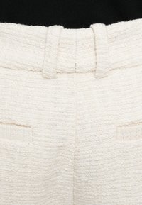 Fehér szövött anyagból készült nadrág, amely texturált felülettel, varrott derékrésszel és két hátsó zsebbel rendelkezik. Minimalista kialakítás tiszta vonalakkal.