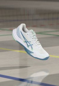 Scarpa da ginnastica bianca con accenti blu e verdi che fluttua sopra un pavimento di campo indoor segnato con linee colorate.