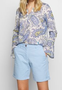 Blouse boutonnée avec un imprimé paisley bleu, jaune et crème ; coupe ample, manches longues. Shorts en coton bleu clair, retroussés avec un détail de bouton.