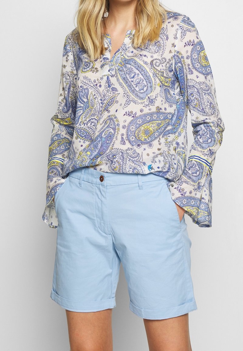 Blouse boutonnée avec un imprimé paisley bleu, jaune et crème ; coupe ample, manches longues. Shorts en coton bleu clair, retroussés avec un détail de bouton.