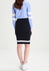 Žena má na sobě černou sukni ke kolenům s dvěma bílými pruhy u lemu, světle modrý top s dlouhými rukávy a bílými pruhy, a bílé tenisky.