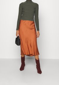 Bruine satijnen midi-rok met een lichte plooi, gecombineerd met een getextureerde donker groene top met lange mouwen. Het model houdt een zwarte handtas vast en draagt bordeauxrode laarzen.