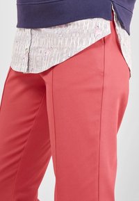 Spodnie w kolorze koralowym wykonane z gładkiej tkaniny, o prostym kroju i widocznych bocznych szwach, zestawione z wzorzystą koszulą pod spodem.