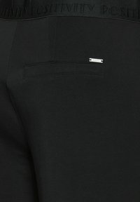 Černé sportovní kalhoty s texturovaným pasem, na kterém je embosovaný nápis "POZITIVITA". Obsahují malý stříbrný kovový logo akcent a zadní kapsu.