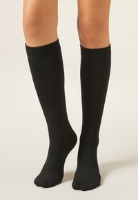 Calzini neri fino al ginocchio in tessuto liscio, con design aderente e polsini a coste, che arrivano appena sotto il ginocchio.