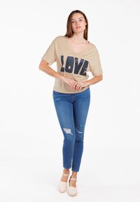 Camiseta beige con la palabra "AMOR" en glitter azul marino, de manga corta, combinada con jeans skinny desgastados azules y espadrilles de color claro.