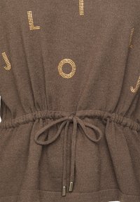 Tissu tricot marron avec lettres cloutées dorées, taille froncée avec ceinture nouée en nœud et embouts métalliques sur les cordons.