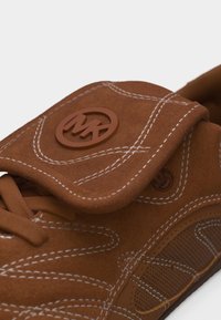 Καφέ σουέντ αθλητικό παπούτσι με λευκή ραφή, αναδιπλούμενο καπάκι, λογότυπο "MK" από καουτσούκ και κορδόνια σε λευκό φόντο.