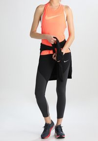 Nike-Sport-Top in leuchtendem Orange mit weißem Logo, kombiniert mit schwarzen, eng anliegenden Leggings mit grauen Akzenten und einer schwarzen Jacke, die um die Taille gebunden ist.