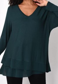 Tealgrön tunika med V-ringning, långa ärmar och lager av volanger längst ner. Tillverkad av mjukt material med en lös passform och en flödande design.