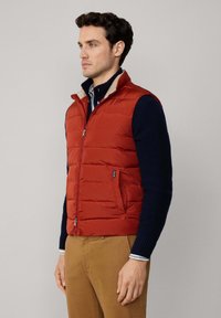 Chaleco inflable rojo con cremallera en la parte delantera, cuello beige y bolsillos laterales, superpuesto sobre un suéter azul marino. Llevado con pantalones caquis.