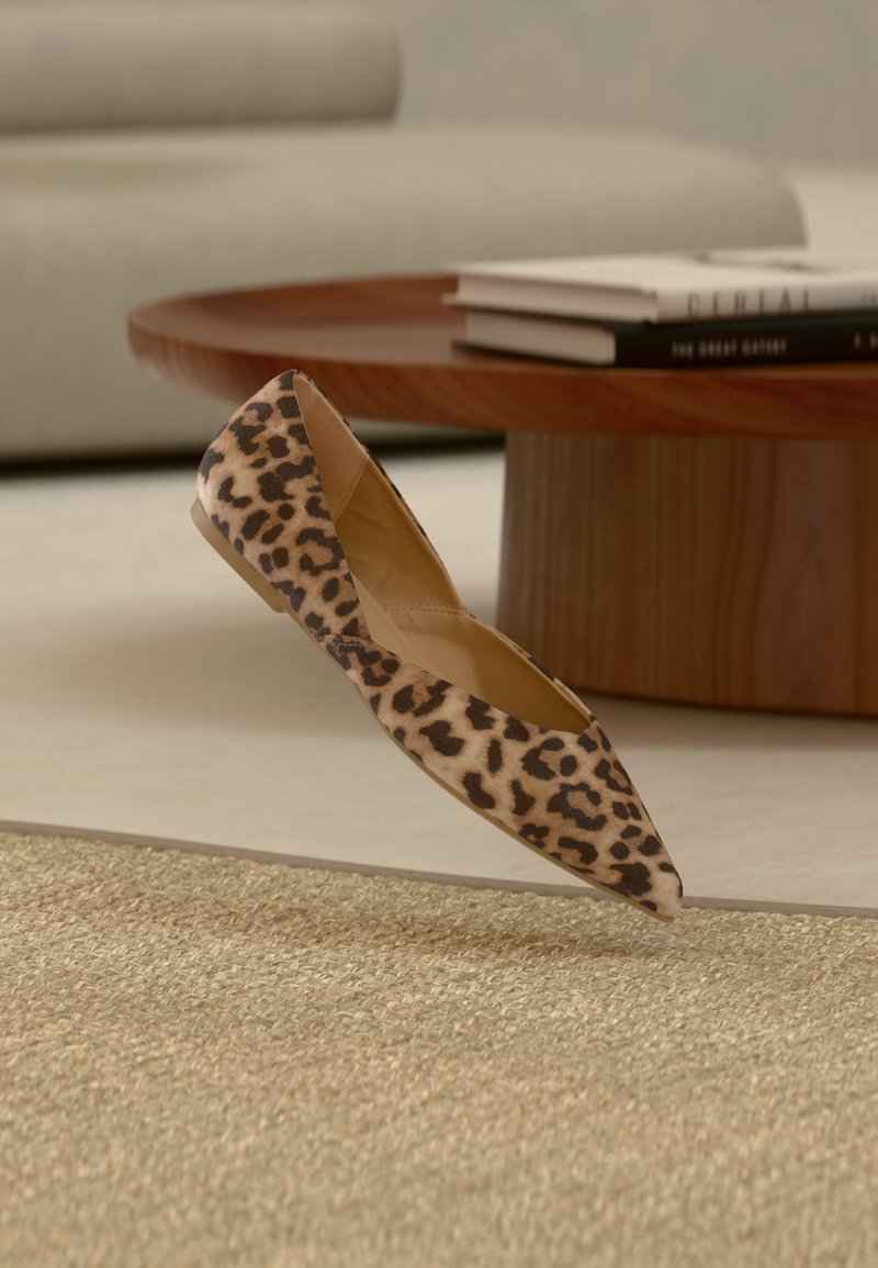 Γυναικείο επίπεδο παπούτσι με λεοπάρ σχέδιο και μυτερή άκρη κοντά σε στρογγυλό ξύλινο τραπεζάκι καφέ με στοιβαγμένα βιβλία πάνω σε μπεζ χαλί και φόντο έναν καναπέ.
