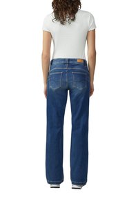 Frau mit lockigem, dunklem Haar, die ein weißes Kurzarmoberteil und blaue, gerade Jeans trägt, steht mit dem Rücken zu einem weißen Hintergrund.