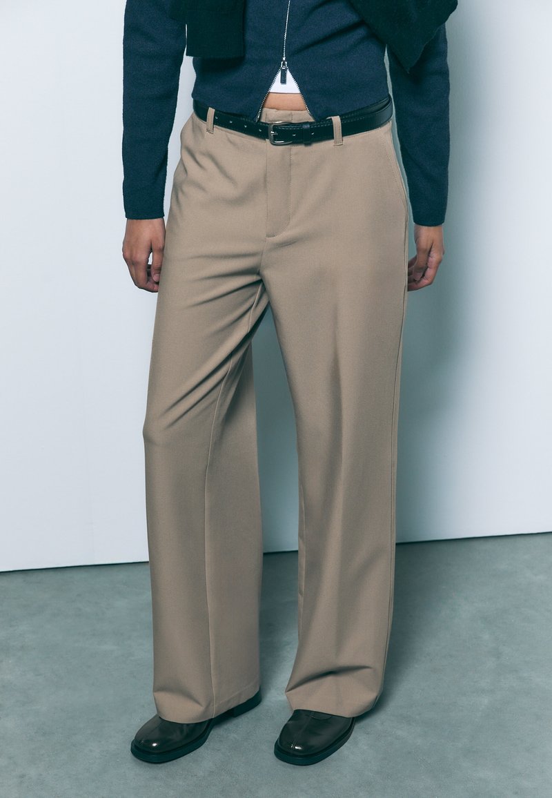 Pantalon beige à jambes larges avec une texture lisse, présentant des plis à l'avant et une ceinture noire. Associé à des chaussures noires brillantes.
