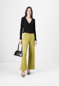 Top avvolgente nero a maniche lunghe con orlo a volant, abbinato a pantaloni gialli a vita alta e a gamba larga con texture. Gli accessori includono una borsa nera e tacchi color taupe.