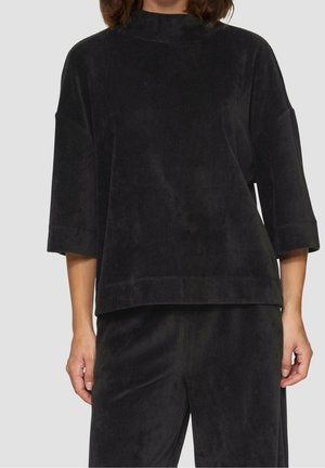 Femme portant un haut noir côtelé ample avec des manches au coude et un pantalon noir côtelé assorti sur un fond neutre.
