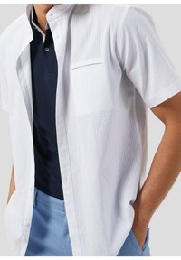 Uomo che indossa una camicia bianca a maniche corte con bottoni sopra un polo blu navy, con pantaloni azzurri e le mani nelle tasche.
