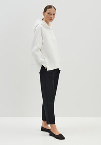 Haut à capuche blanc oversize avec une poche avant, associé à un pantalon noir slim et des ballerines noires, se tenant sur un fond uni.