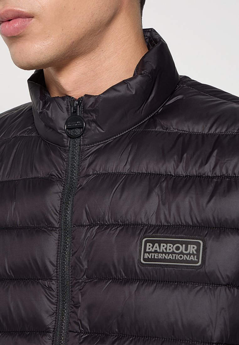 Schwarze Steppjacke mit hohem Kragen, durchgehendem Reißverschluss und Barbour International Logo-Patch. Glatte Textur mit Steppmuster.