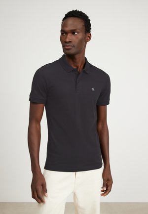 Black polo shirt made from smooth fabric, featuring a classic collar, short sleeves, and a small logo on the left chest. Paired with light trousers.
