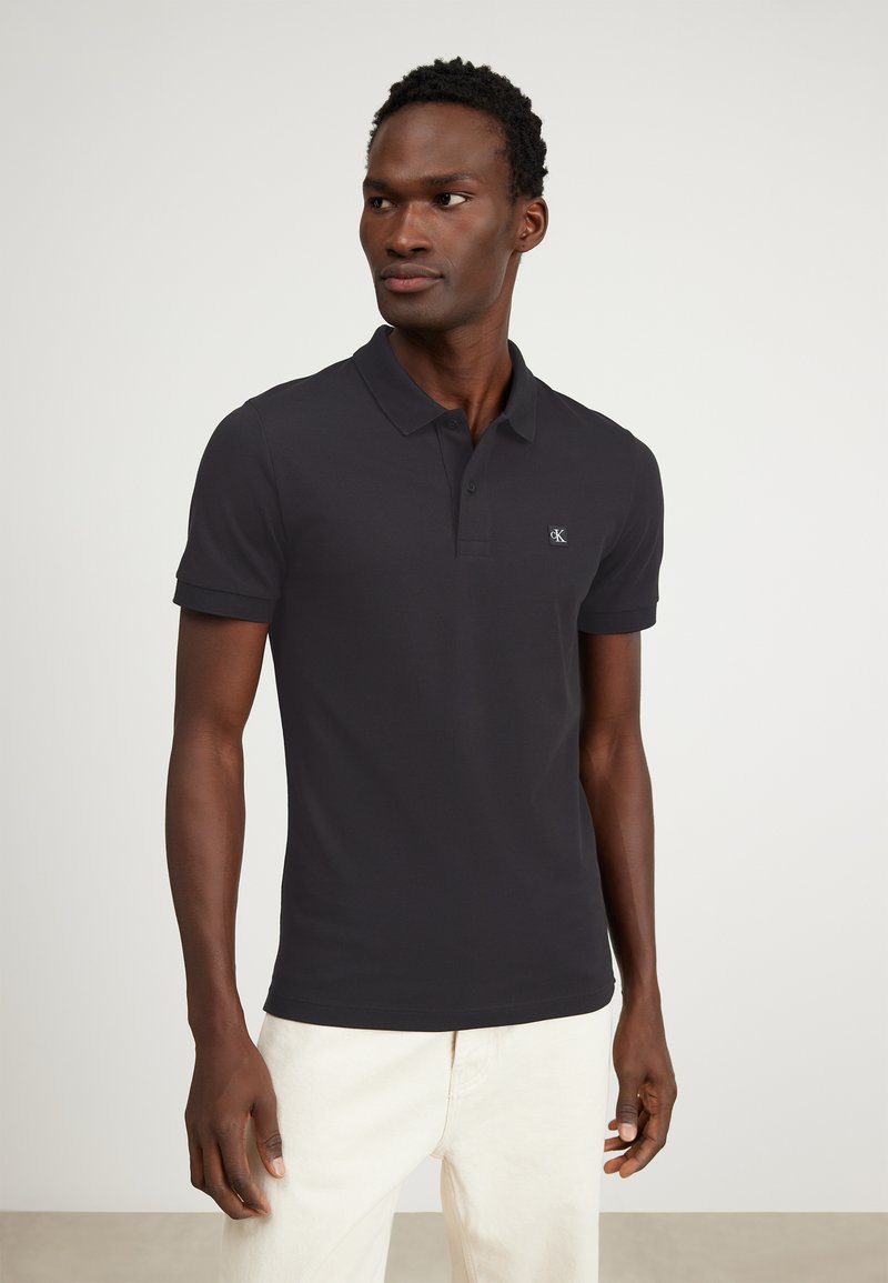 Camisa polo negra hecha de tejido suave, con cuello clásico, mangas cortas y un pequeño logo en el lado izquierdo del pecho. Combinada con pantalones claros.