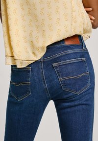 Jeans de mezclilla azul oscuro con costuras amarillas, que cuentan con dos bolsillos traseros y una etiqueta de cuero en la cintura. La tela parece suave y ajustada.
