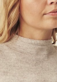 Világos bézs pulóver magas nyakkivágással, bordás díszítéssel. Puha textúra, finom eltérések, és kényelmes illeszkedés a nyak körül.