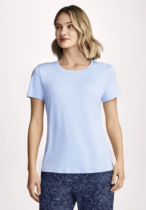 Frau mit schulterlangen blondem Haar, die ein hellblaues T-Shirt mit kurzen Ärmeln und dunkelblaue, gemusterte Hose trägt, steht und schaut nach rechts.