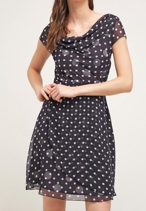 Femme portant une robe noire longueur genou à pois blancs, manches courtes et encolure drapée, les mains jointes à la taille.