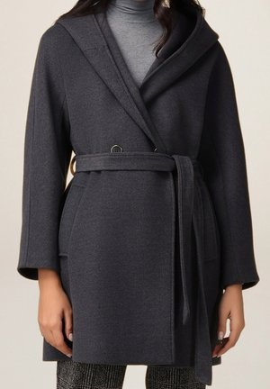 Femme portant un manteau ceinturé gris foncé sur un pull à col roulé gris clair, vue de face avec manches longues et poches latérales.