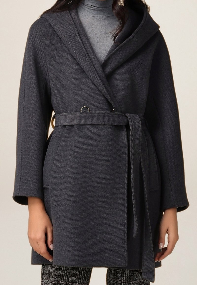 Femme portant un manteau ceinturé gris foncé sur un pull à col roulé gris clair, vue de face avec manches longues et poches latérales.