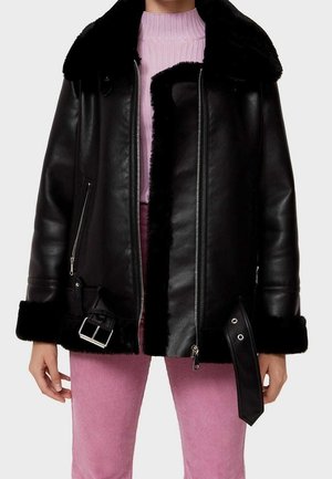 Zwarte leren jas met bontvoering, ritssluiting voor, gespsluiting, gedragen over een roze geribde trui en roze broek.