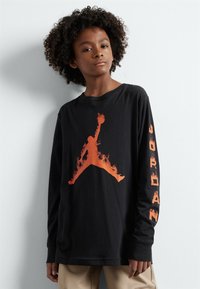 Camiseta de manga larga negra con un gran logo de Jumpman con patrón de llamas naranjas en el frontal y la palabra "JORDAN" impresa en naranja a lo largo de la manga.