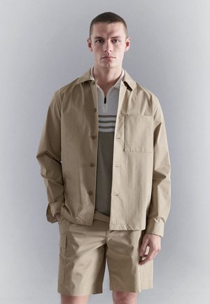 Joven con camisa beige abotonada y pantalones cortos a juego, de pie frente a un fondo liso de color gris claro, mirando hacia adelante.