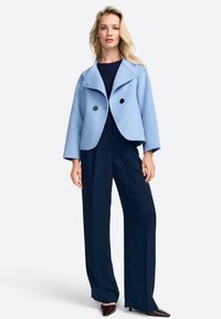 Giacca cropped azzurra con colletto a scialle, due bottoni neri, abbinata a pantaloni a gamba larga blu navy e scarpe con punta.